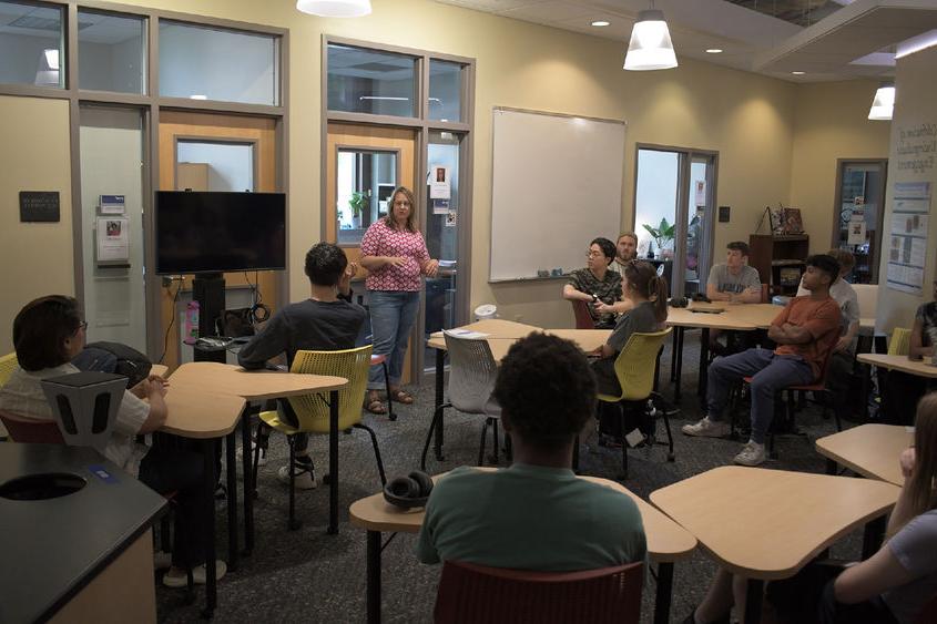 Person stands at the front of a room, talking to other people sitting at tables. 